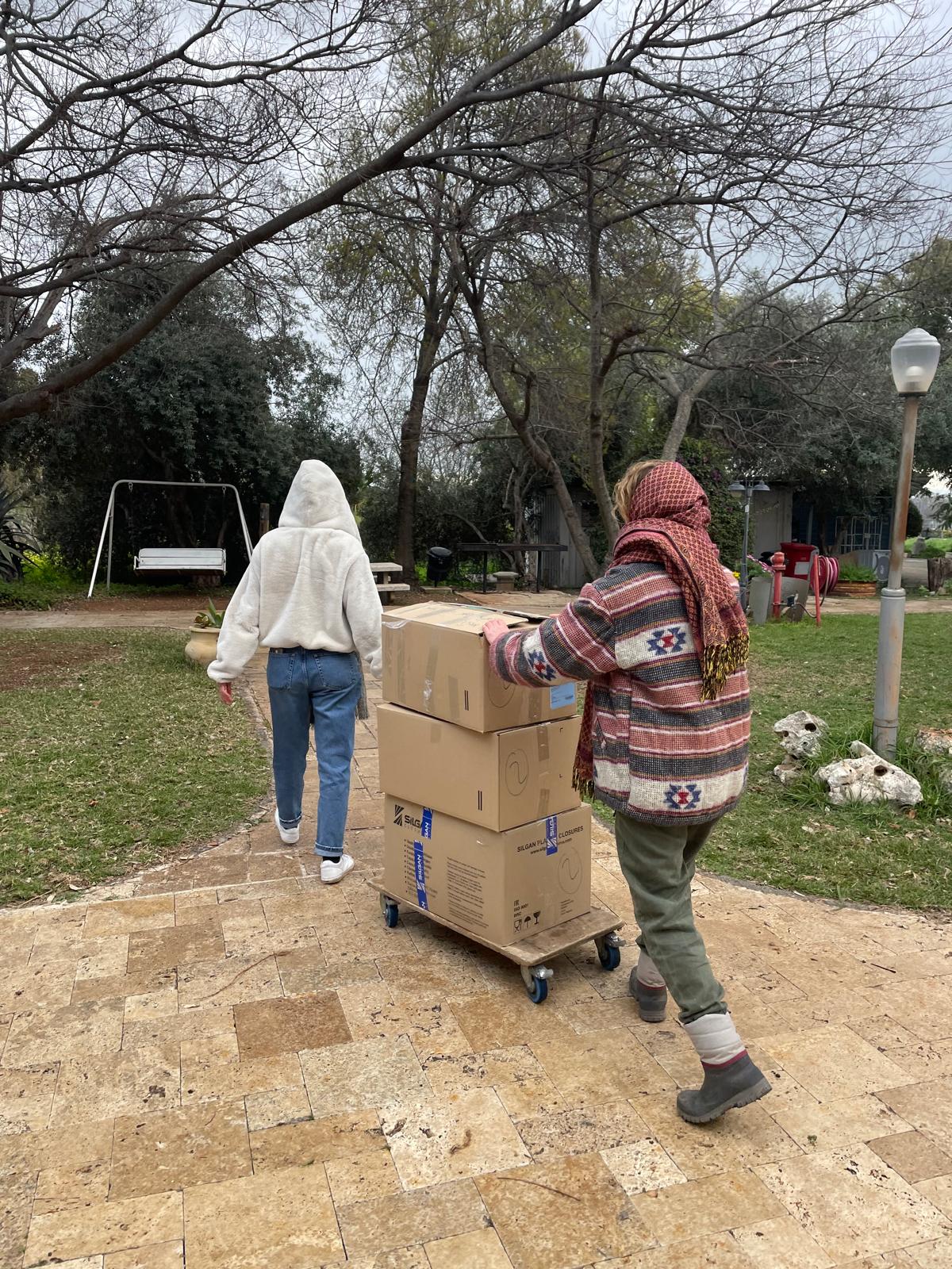 שני אנשים מובילים קרטונים על עגלה בגינה פתוחה עם עצים ודשא.