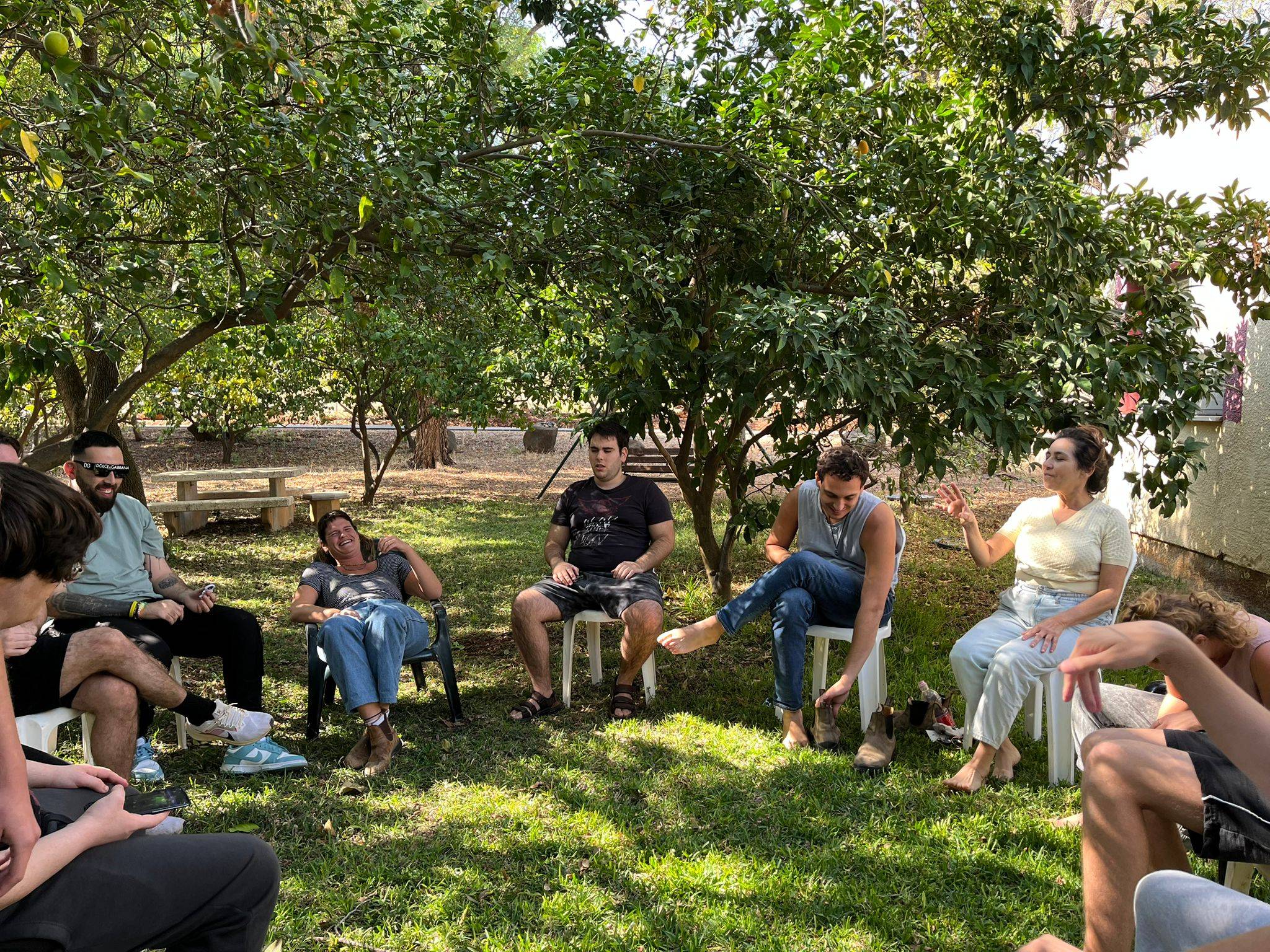 קבוצה יושבת במעגל מתחת לעצים בפארק, מדברים ומחייכים. אווירה נינוחה וירוקה.