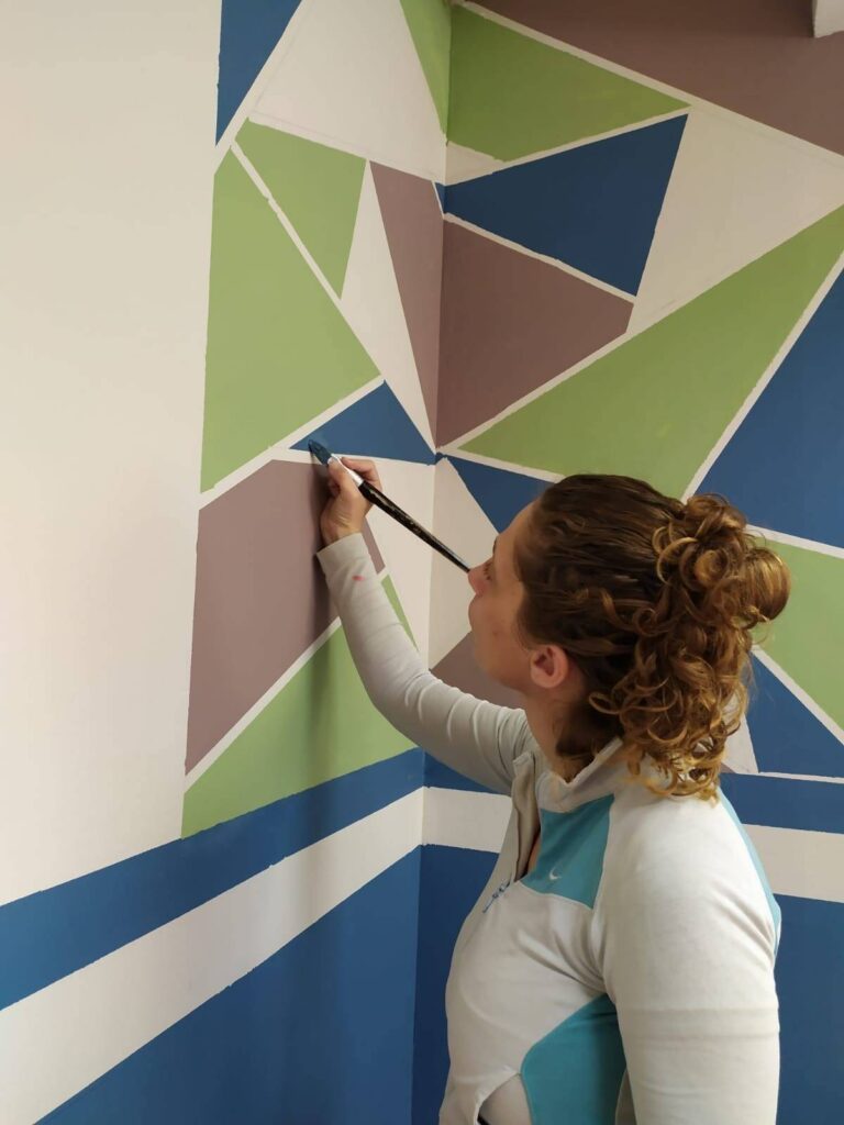 אדם מצייר קיר בעזרת צבעים כחול כחלק מהתרחבות העמותה לחיפה, ירוק וסגול בעיצוב גאומטרי.