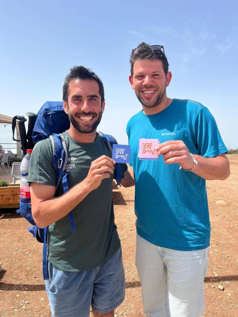 שני אנשים מחזיקים כרטיסים צבעוניים בחוץ ביום שמש, אחד מהם עם תיק גב על השביל.