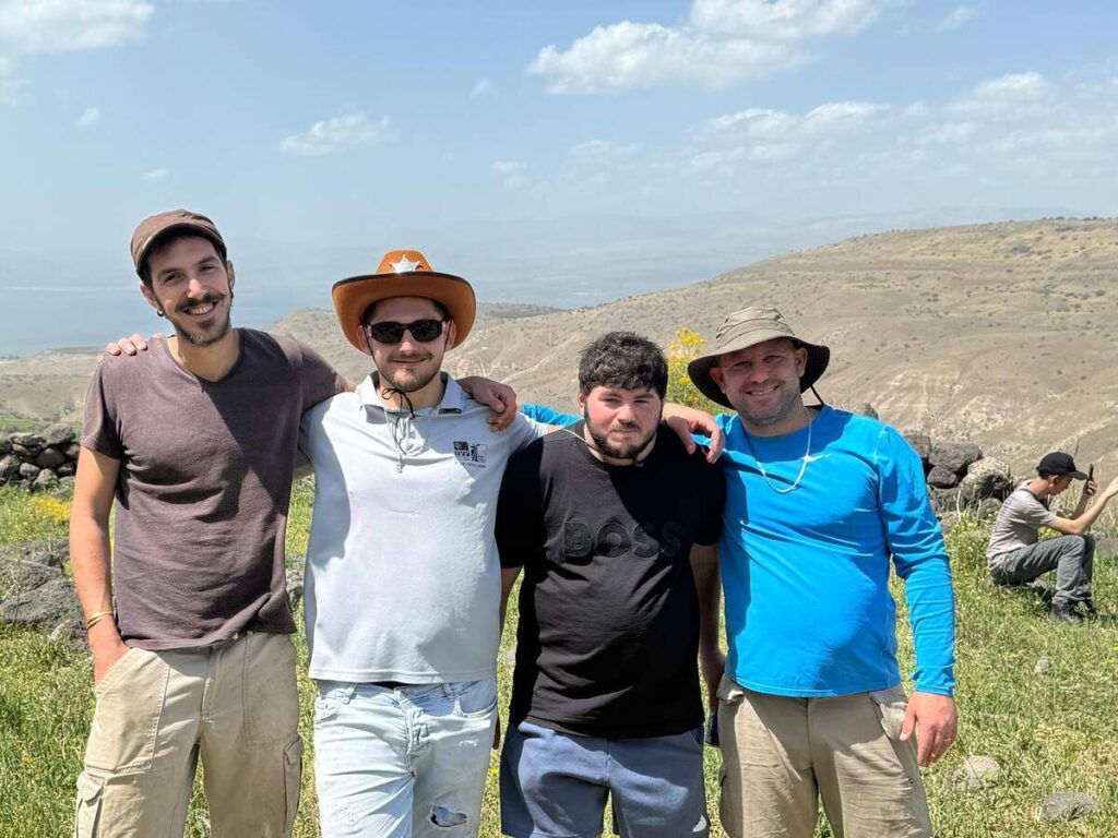 ארבעה אנשים מחייכים בטיול בנוף הררי, אחד מהם חובש כובע בוקרים. ברקע ים והרים.
