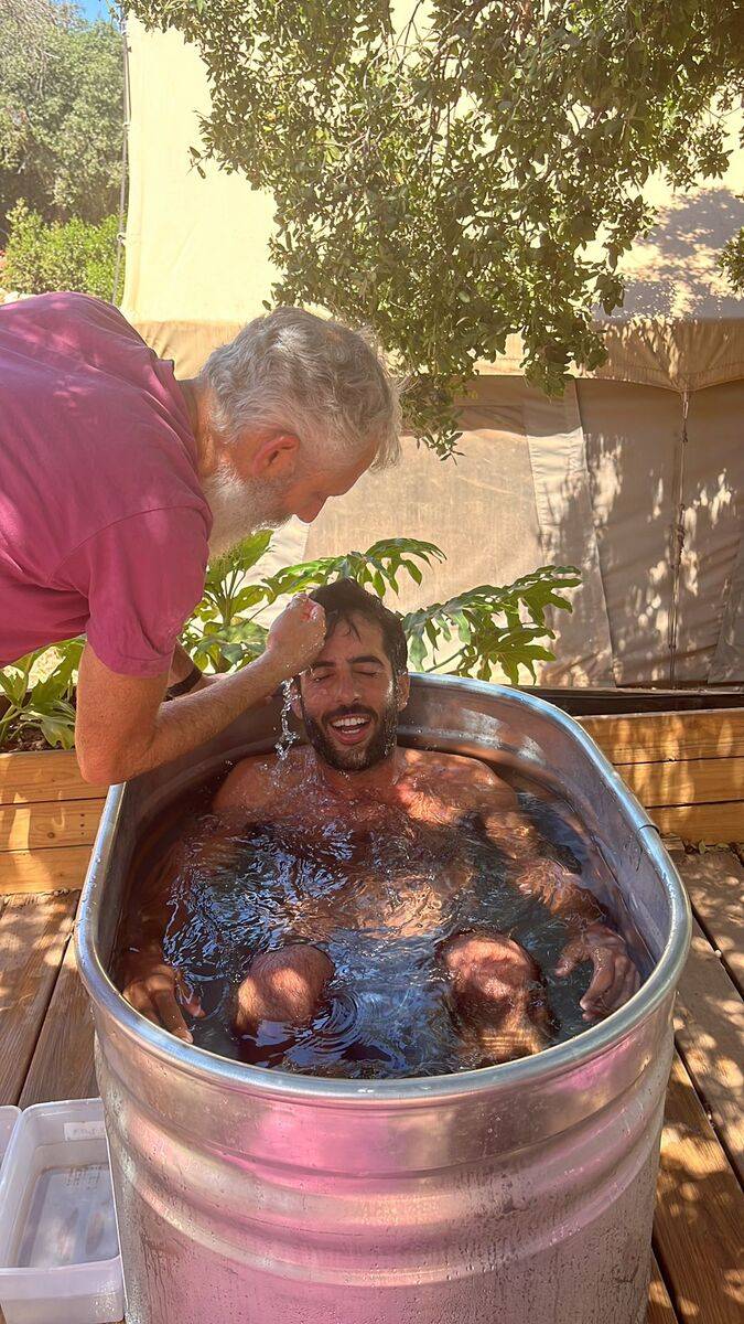 שני אנשים בחיק הטבע, אחד יושב באמבט מתכת ומעליו מוזגים מים, עץ ברקע.