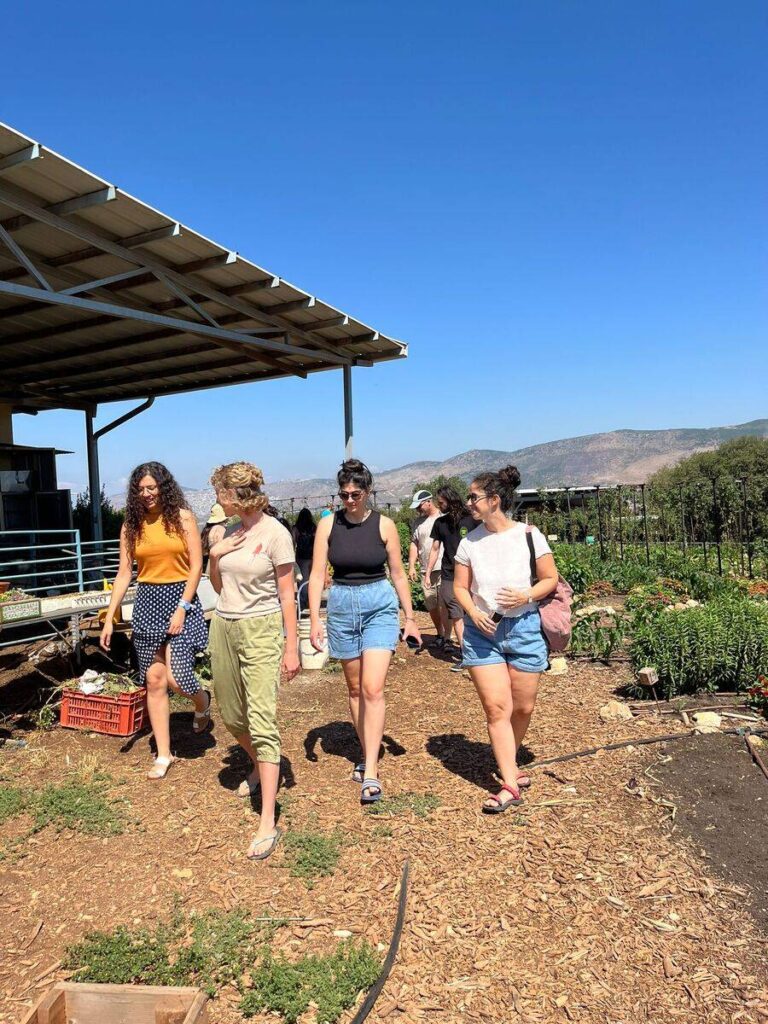 קבוצת אנשים מטיילת בטבע ליד גן ירק ומחסן, נוף הרים ברקע ויום עם שמיים כחולים.