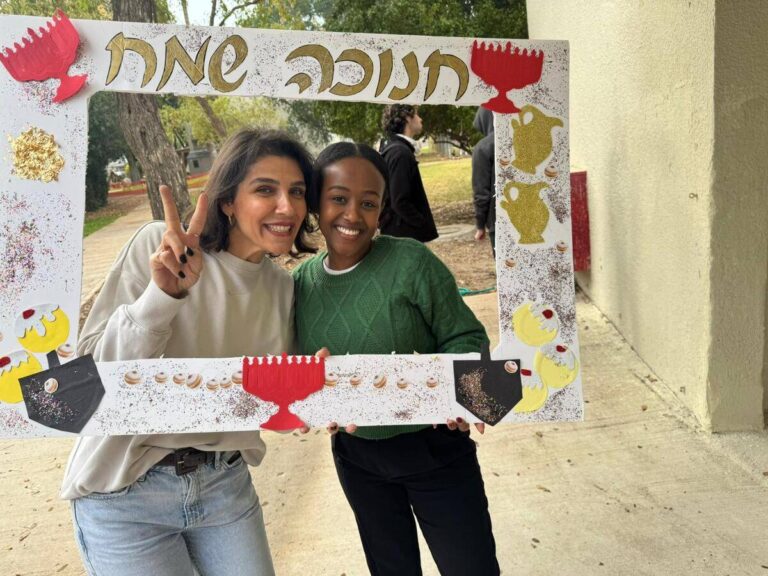 שתי נשים מחזיקות מסגרת עם כיתוב חנוכה שמח בחיוך, ברקע נוף חיצוני ירוק.