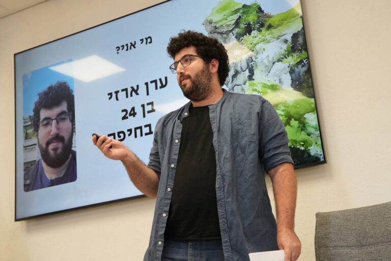 אדם מציג מול מסך עם תמונה וטקסט בעברית, בחדר ישיבות.