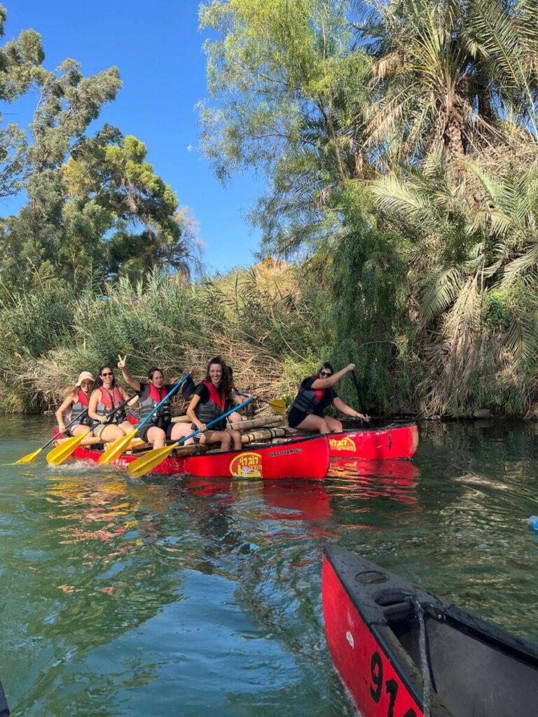 אנשים בחותרים בקיאקים אדומים בנהר עם נוף עצים ירוקים ושמים כחולים.